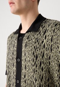 Knit shirt with a black collar, featuring a green and black pattern of elongated shapes. Buttons down the front; short sleeves.