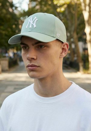 Junger Mann mit einer hellgrünen New York Yankees Baseballkappe und weißem Hemd, stehend im Freien in einem von Bäumen gesäumten städtischen Gebiet.