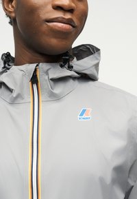 Κοντινό πλάνο ενός ατόμου που φοράει ένα ανοιχτό γκρι μπουφάν με κουκούλα και εμπρός φερμουάρ με κίτρινες, λευκές και ναυτικές ρίγες, καθώς και το λογότυπο K-Way.