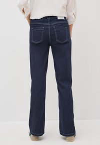 Jeans évasés bleu foncé à taille moyenne, avec des poches arrière et des coutures blanches contrastantes ; portés avec un haut de couleur claire.