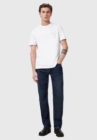 T-shirt bianca in cotone con collo rotondo, abbinata a jeans denim blu scuro. Il modello indossa scarpe nere. Design semplice, senza motivi o loghi visibili.