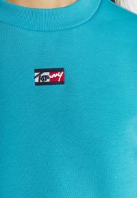 Turkos munkjacka med rund halsringning som har en broderad logotyp i röd, vit och svart. Tyget är mjukt och strukturerat.