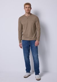 Langärmliges braunes Henley-Shirt mit Knopfblende, kombiniert mit blauen Jeans und weißen Sneakers. Einfaches Design, weiche Textur und lässiger Look.