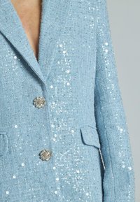 Blazer azzurro chiaro con texture e decorazioni in paillettes, dotato di un bottone floreale, rever a punta e due tasche frontali.