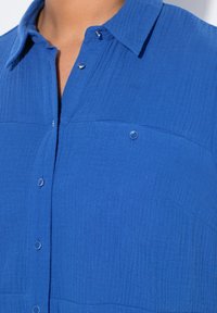 Camicia azzurra con bottoni, caratterizzata da un tessuto testurizzato, due tasche sul petto e bottoni in tono argentato. Il colletto ha un design classico.