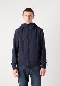 Marineblauwe capuchonjack van lichtgewicht stof, met een ritssluiting aan de voorkant, elastische manchetten en een ontspannen pasvorm. Gecombineerd met donkere denim jeans.