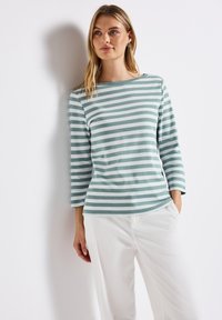 Gestreepte lange mouw top in licht teal en wit, gemaakt van zachte stof met een bootnek en een ontspannen pasvorm. Gecombineerd met witte pants.