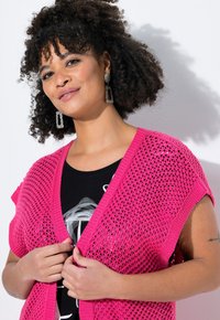 Rosa gestrickte Kimono-Cardigan mit kurzen Ärmeln und offener Vorderseite, ausgestattet mit einer lockeren Webstruktur. Darunter ein schwarzes Graphic-T-Shirt.