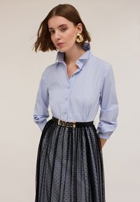 Cămașă cu dungi albastre și albe, cu guler înalt, asortată cu o fustă texturată neagră și un curea neagră lucioasă dotată cu accesorii din aur.