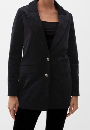 Femme portant un blazer noir cintré avec des revers crantés, deux boutons sur le devant et des poches à rabat, par-dessus un top noir sur un fond uni.