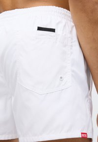 Valged ujumispüksid, mis on valmistatud kergest kangast, varustatud tagataskuga, millel on must rihm ja väikese punase logomärgiga ääres.