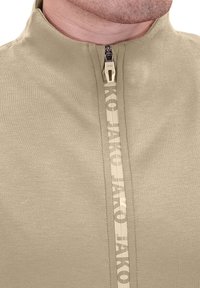 Pullover a collo alto in tessuto khaki chiaro con zip frontale, dotato di un tirazip con logo e una superficie liscia e opaca.