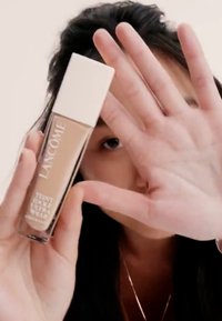 Base de Lancôme en un frasco de vidrio transparente con tapa blanca; líquido beige claro visible. La mano de un modelo cubre parcialmente el producto.