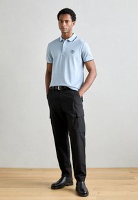 Camisa polo azul claro con cuello negro y emblema, combinada con pantalones cargo negros y botas negras hasta el tobillo, sobre un fondo neutro.