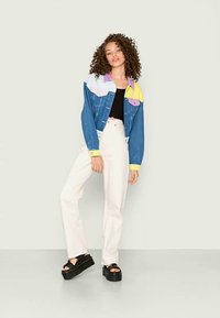 Jaqueta de ganga com um design com blocos de cor em azul, roxo e amarelo, combinado com uma blusa preta e calças brancas de cintura alta. Sandálias de plataforma pretas.
