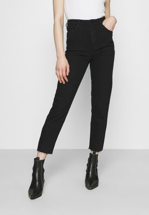 Osoba nosící černé džíny s vysokým pasem a neupravenými lemy, černé kotníkové boty a bílý tílkový top, stojící před čistým pozadím.