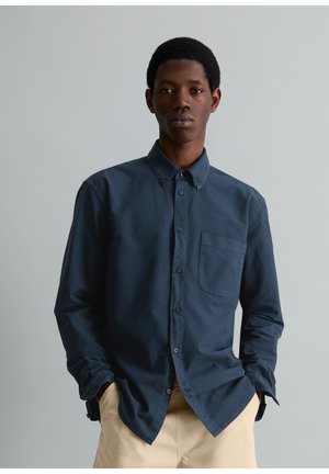 Homme aux cheveux courts portant une chemise boutonnée bleu foncé et un pantalon beige, debout devant un fond gris clair uni.