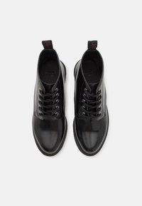 Botas de tobillo de cuero negro con punta redonda, acabado brillante, seis ojales de metal y cordones negros. Suela de goma con detalle texturizado y tira de agarre.