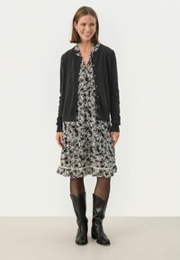 Zwarte cardigan, bloemenprint jurk in marineblauw en cr�ème met ruches aan de onderkant, zwarte panty, zwart leren laarzen tot de knie. Model staat voor een witte achtergrond.