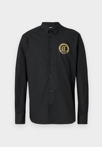 Camisa negra de botones con un logo circular dorado en el lado izquierdo del pecho. Hecha de tela suave con mangas largas y un diseño clásico de cuello.
