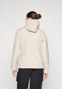 Veste beige clair avec capuche, fabriquée en tissu doux. Elle présente une coupe ajustée, des manches longues et des détails de couture visibles.