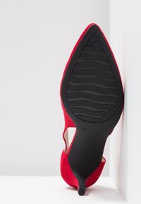 Chaussure à talon haut pointue en daim rouge positionnée verticalement, montrant la semelle noire avec motif de bande de roulement et indication de la taille 37.