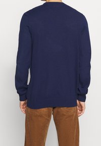 Marineblauer Strickpullover mit rundem Ausschnitt, gerippten Bündchen und Saum. Mit Abnähern auf der Rückseite und glattem Material. Kombiniert mit braunen Cordhosen.