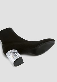 Bottine noire avec un dessus élastique, dotée d'un talon bloc transparent et d'une semelle texturée pour une bonne adhérence. La semelle présente des rainures horizontales.