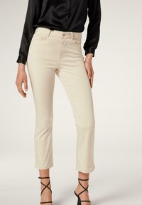 Béžové bavlněné kalhoty z látky s příměsí, zkráceného střihu a přiléhavého designu, se zapínáním na knoflík vpředu a bočními kapsami. V kombinaci s černou saténovou blůzou.