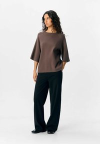 Haut marron à manches courtes avec une coupe décontractée et un col rond, associé à un pantalon large noir et des chaussures noires, sur un fond neutre.