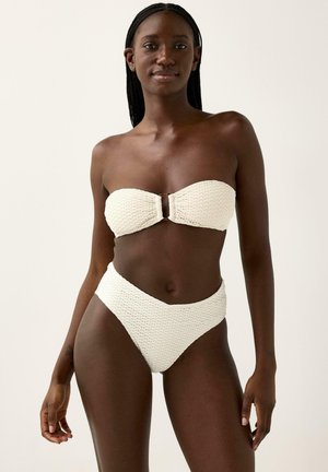 Kvinde med mørk hud iført en tekstureret hvid, stropfri bandeau-bikinitop og bikinitrusser med høj talje, stående mod en ensfarvet baggrund.