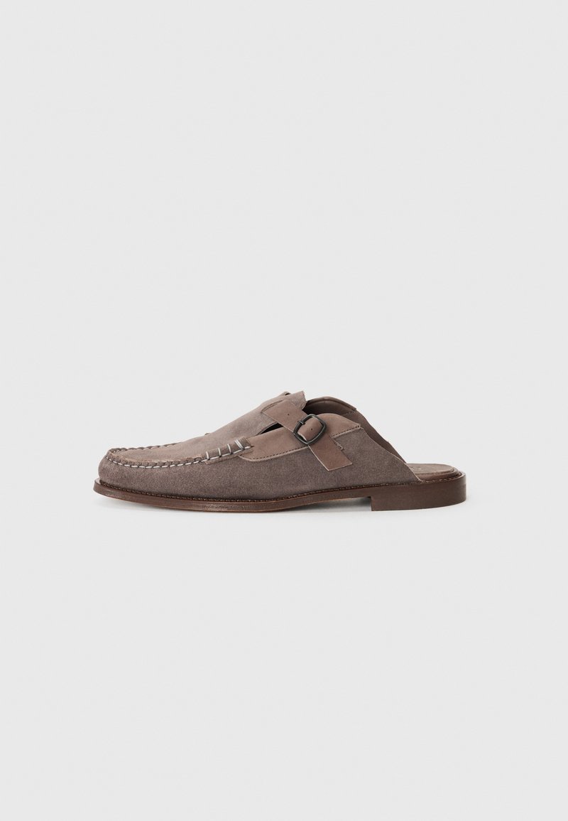 Scarpe slip-on in suede marrone con punta arrotondata, cuciture decorative e fibbia laterale. Suola piatta in cuoio e tomaia strutturata.