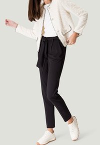 Biała tweedowa kurtka z metalicznymi akcentami, noszona na białej bluzce. Czarna dopasowana spodnie i białe sneakersy z teksturowaną podeszwą.