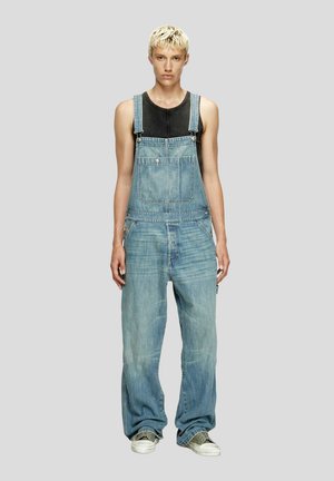 Salopette di denim blu con bretelle regolabili, tasca frontale e gambe larghe, indossata sopra una canottiera nera. Scarpe da ginnastica completano l'outfit.