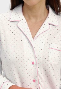 Camisa de piyama blanca con pequeños patrones de corazones coloridos en rosa, marrón y gris claro. Presenta un bolsillo, botones rosas y un ribete en contraste.