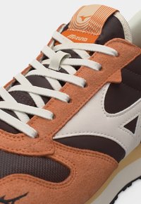 Κοντινή λήψη καφέ, λευκού και πορτοκαλί αθλητικού παπουτσιού Mizuno με λευκά κορδόνια, πάνελ από σουέντ και δίχτυ, και λεπτομέρεια με το λογότυπο στη γλώσσα.