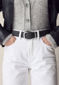 Osoba ubrana w białe spodnie z czarnym tłoczonym skórzanym paskiem, szary rozpinany sweter z miękkiego materiału oraz czarną skórzaną kurtkę, trzymająca ręce w kieszeniach.