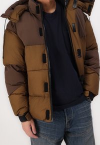 Braune und beige Steppjacke mit einem Reißverschluss vorne, elastischen Bündchen und schwarzen Akzenten. Der Stoff wirkt glatt mit einer gesteppten Textur.