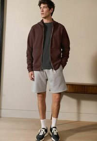 Maroone Zip-Hoodie, graues T-Shirt und graue Shorts. Schwarze Sneaker mit weißen Sohlen und gemusterten Socken. Glatter Stoff, lässiges Design.