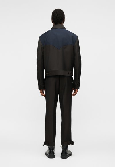 Άνδρας με την πλάτη γυρισμένη φοράει μαύρο παντελόνι, μαύρες μπότες και μαύρο μπουφάν με σκούρο μπλε τμήμα στην πάνω πλάτη και λεπτομέρειες με κουμπιά.