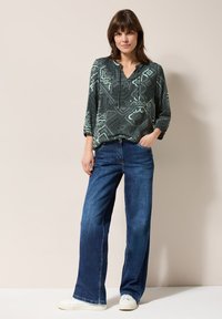 Blouse vert foncé à motifs avec des manches trois-quarts, assortie à un jean large bleu et des baskets blanches. Texture de tissu lisse et coupe décontractée.