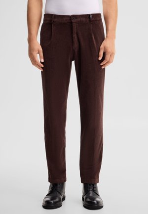 Uomo che indossa pantaloni di velluto a coste marrone scuro con tasche laterali e scarpe stringate in pelle nera su sfondo semplice.