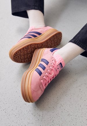Person iført lyserøde Adidas-sneakers med navy striber og tykke gummsåler, hvide sokker og sorte bukser på en lys plettet gulv.