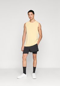 Ανοιχτό κίτρινο αμάνικο πουκάμισο σε συνδυασμό με μαύρο σορτς, λευκά αθλητικά παπούτσια και μαύρες κάλτσες. Το πουκάμισο έχει χαλαρή εφαρμογή και διακριτική λεπτομέρεια με λογότυπο.