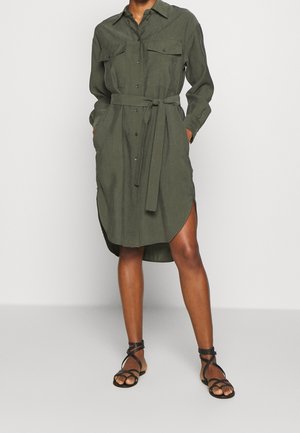Kvinde iført en olivengrøn skjortekjole med bindebånd i taljen og sorte flade sandaler med stropper, stående mod en hvid baggrund.