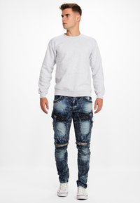 Jeune homme portant un sweatshirt gris clair, un jean bleu usé avec des clous et des zips, et des baskets blanches, se tenant devant un fond blanc.