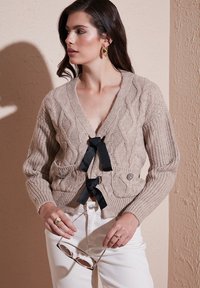 Beige cable-knit cardigan with a deep V-neck, black ribbon ties, and a decorative button. Model holds sunglasses and wears white jeans.
