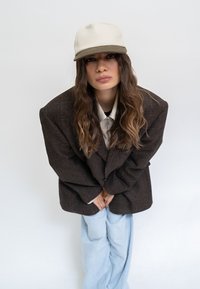 Brauner übergroßer Blazer, weißes Hemd, hellblaue weite Hose, cremefarbene Mütze mit brauem Schirm. Modell posiert mit gefalteten Händen, glattes Haar.