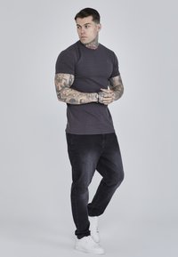Donkergrijze slim fit t-shirt met korte mouwen en een klein logo; gecombineerd met een slim zwarte jeans en witte sneakers. Tatoeages zichtbaar op de armen.