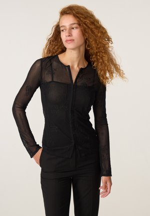 Jonge vrouw met lang krullend haar, gekleed in een zwarte doorschijnende top met lange mouwen en zwarte broek, kijkt iets naar de zijkant.